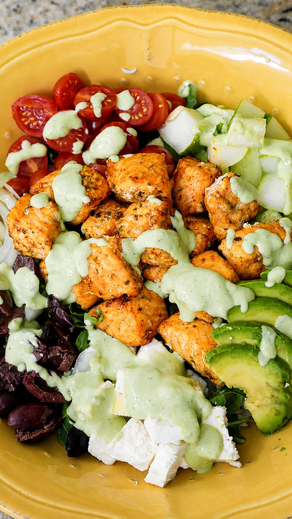Air fryer steelhead bites in a yellow bowl with cherry tomatoes, kalamata olives, feta, avocado and cucumber, drizzled with green goddess tzatziki