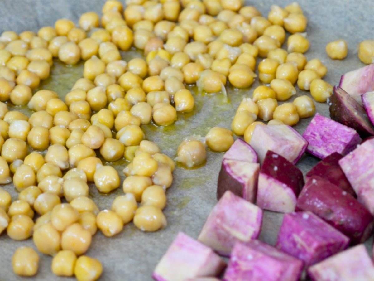 Cubed purple Okinawan sweet potatoes and drained chickpeas tossed with olive oil on a parchment-lined sheet pan, ready to roast