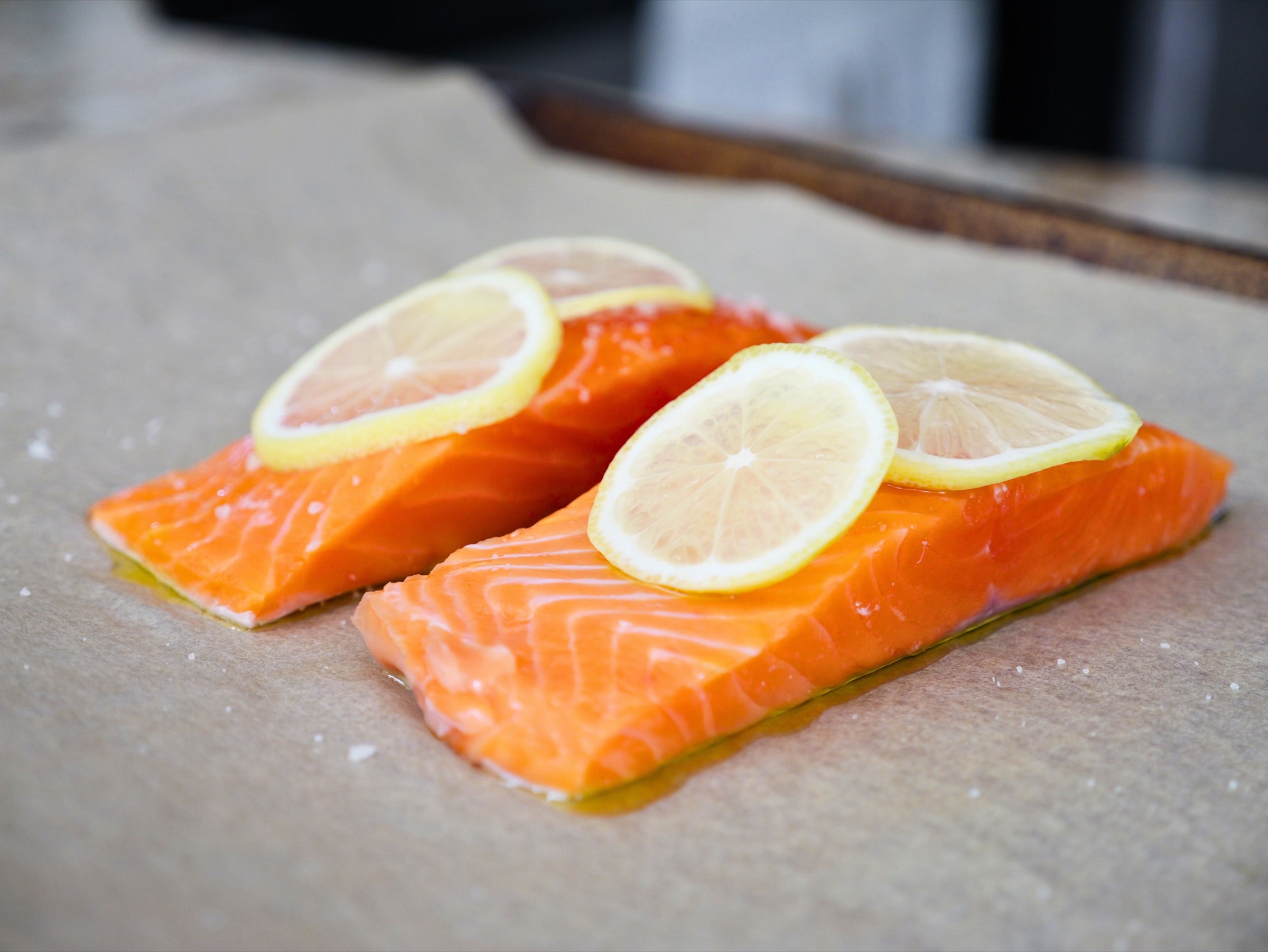 Two raw Seatopia salmon fillets on parchment, drizzled with olive oil, salted and topped with thin lemon rounds before baking