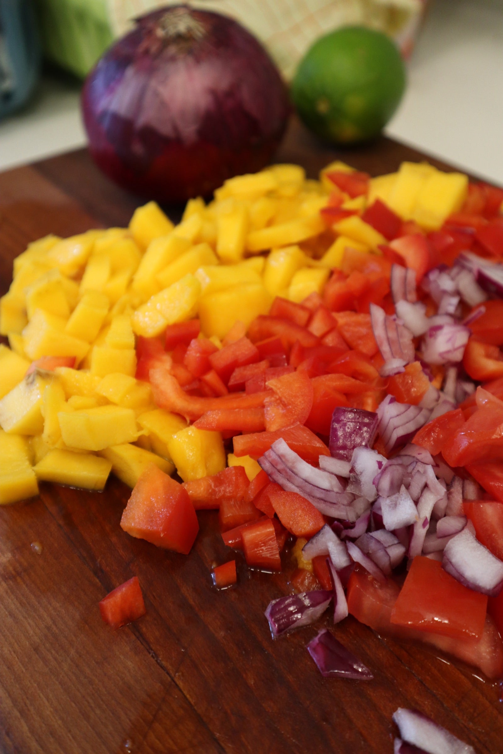 Diced mango, red bell pepper, tomato and red onion on a wooden cutting board for mango salsa