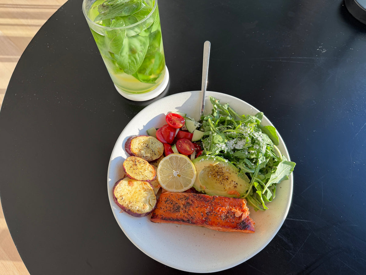 Overhead shot of Kayla Dunham's hot honey King Salmon bowl with air fryer Japanese sweet potato rounds, cucumber and tomato salad, avocado, and greens, beside a glass of basil water