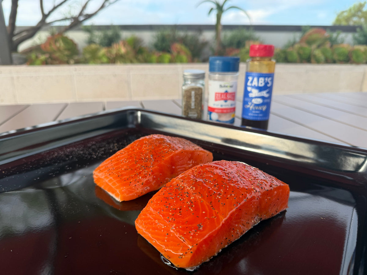 Two Seatopia King Salmon portions seasoned with sea salt and cracked black pepper on a sheet pan, with hot honey and Real Salt in the background