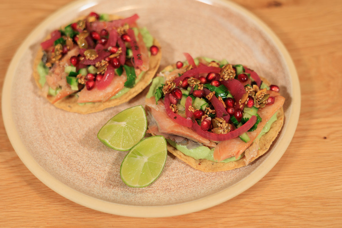 Two smoked trout tostadas topped with cucumber salsa, pickled red onion, pomegranate and salsa macha, with lime wedges on a stoneware plate