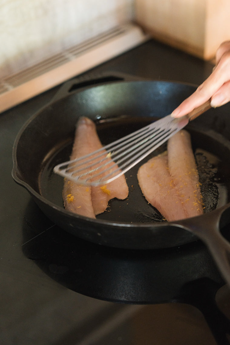 A fish spatula pressing branzino fillets skin-side down in a cast-iron skillet