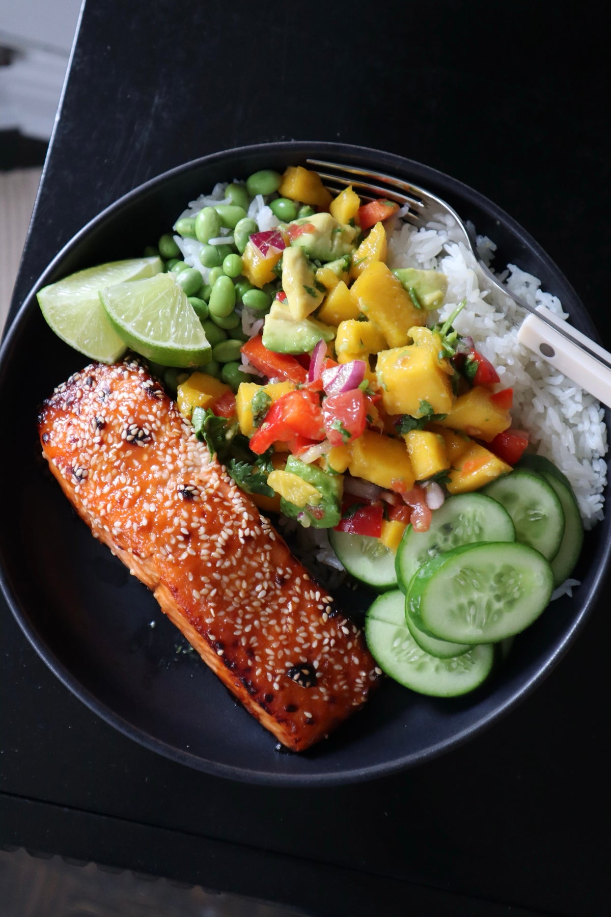 Hot honey salmon bowl with coconut rice, mango salsa, edamame and cucumber in a dark bowl