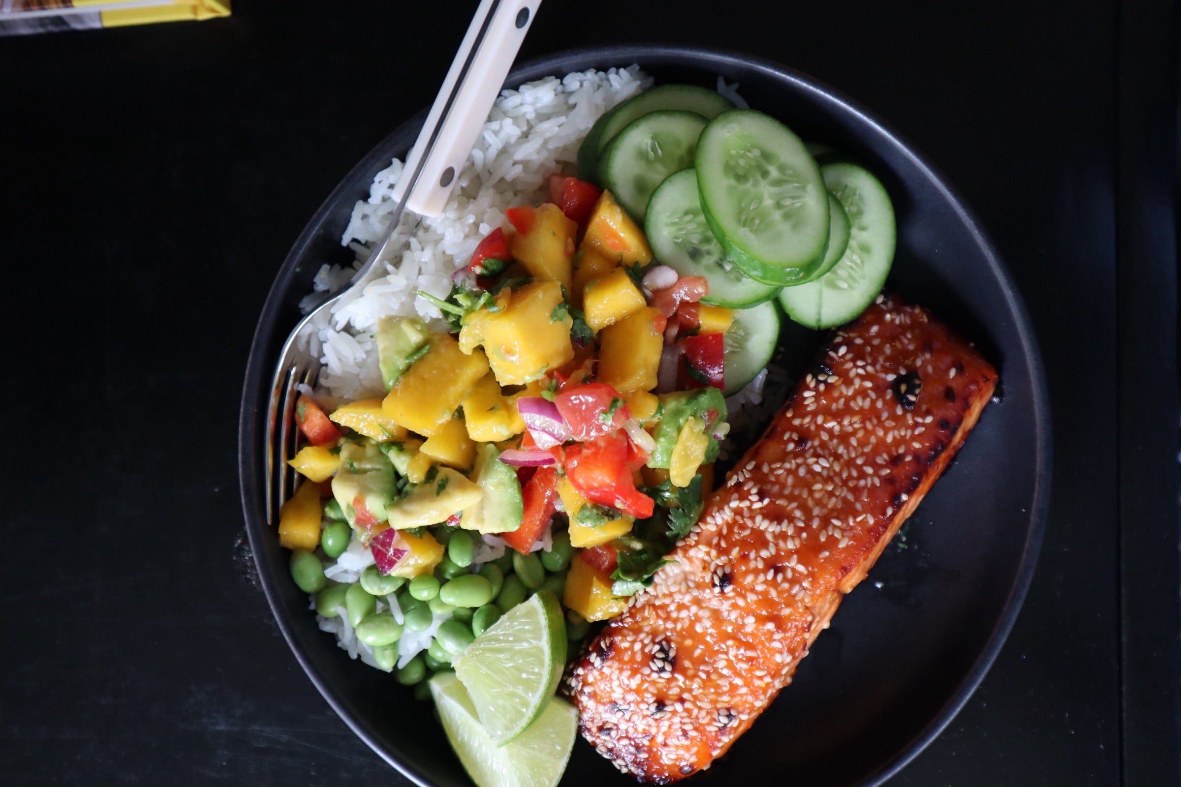 Overhead view of a hot honey salmon bowl with coconut rice, mango salsa, edamame, cucumber and lime