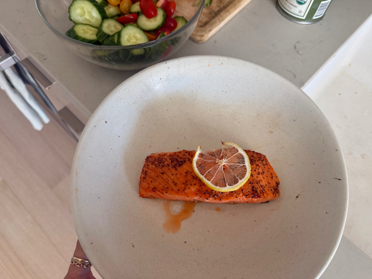 A caramelized hot honey King Salmon fillet on a ceramic plate with a lemon slice, cucumber and tomato salad behind it