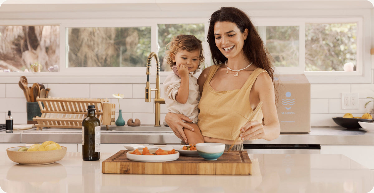 A mother holding her toddler in a bright kitchen preparing seafood, with a Seatopia box on the counter