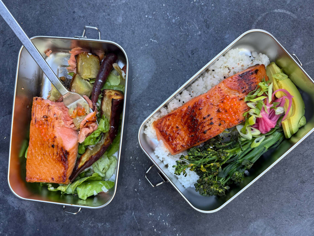 Two stainless bento boxes being built with sushi rice, broccolini, avocado, pickled red onion and a fillet of miso-glazed king salmon