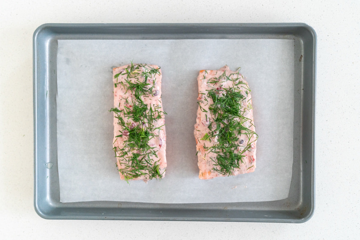 Two Atlantic Salmon portions coated in lingonberry Dijon sauce and dill on a lined sheet pan