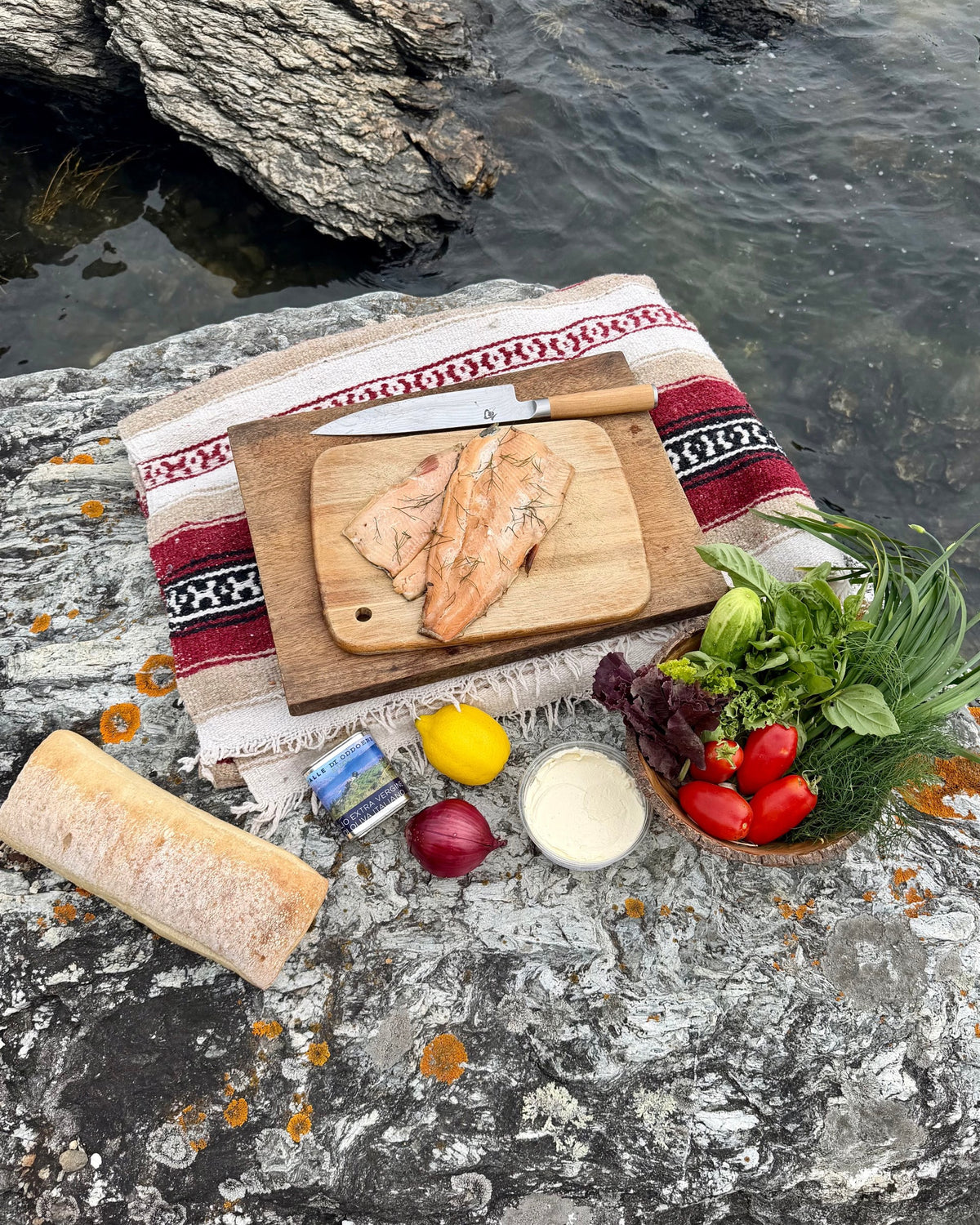 Picnic mise en place on a rock by the water: smoked trout on a board, sourdough loaf, lemon, soft cheese, tomatoes and just-picked herbs