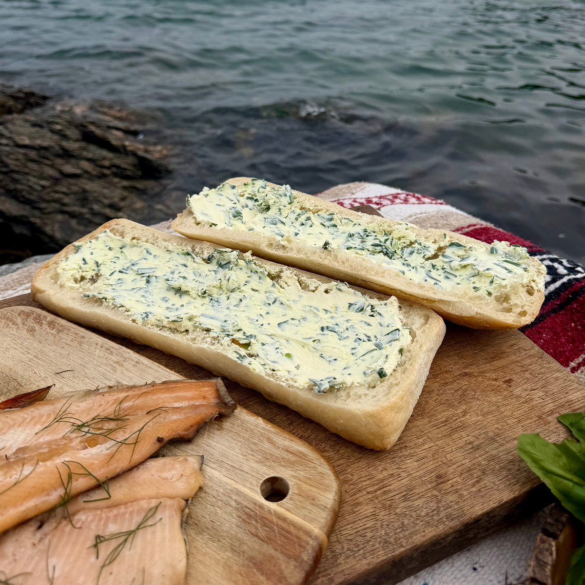 A sourdough loaf split lengthwise and hollowed out, both halves spread with garlic scape and soft cheese, smoked trout fillets on a board alongside
