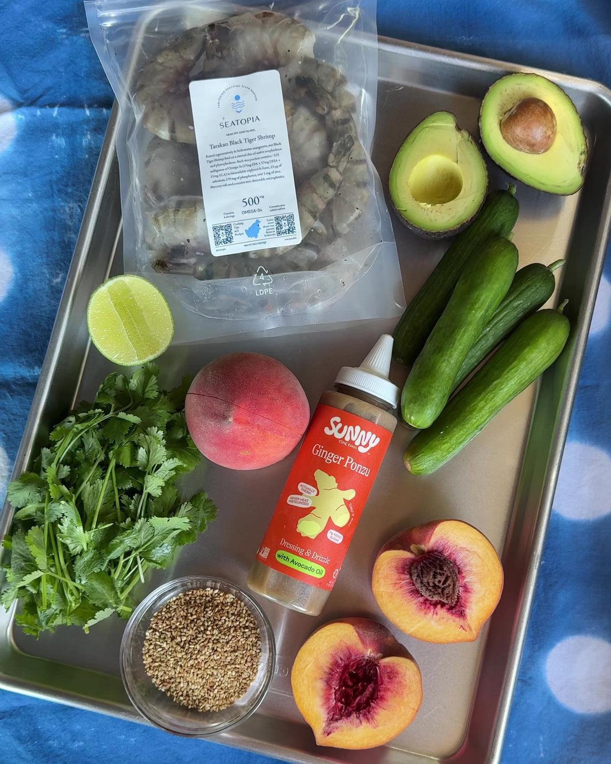 Ingredients for the summery shrimp salad on a sheet pan: a bag of Seatopia Tarakan Black Tiger Shrimp, avocado, Persian cucumbers, peaches, cilantro, toasted sesame seeds, lime and a bottle of Sunny Fine Foods Ginger Ponzu